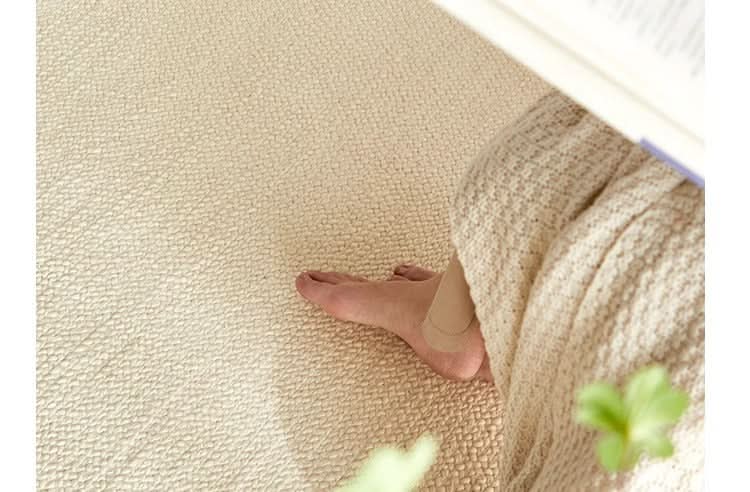 Close-up of a beige braided wool rug and a foot resting under a cozy blanket.